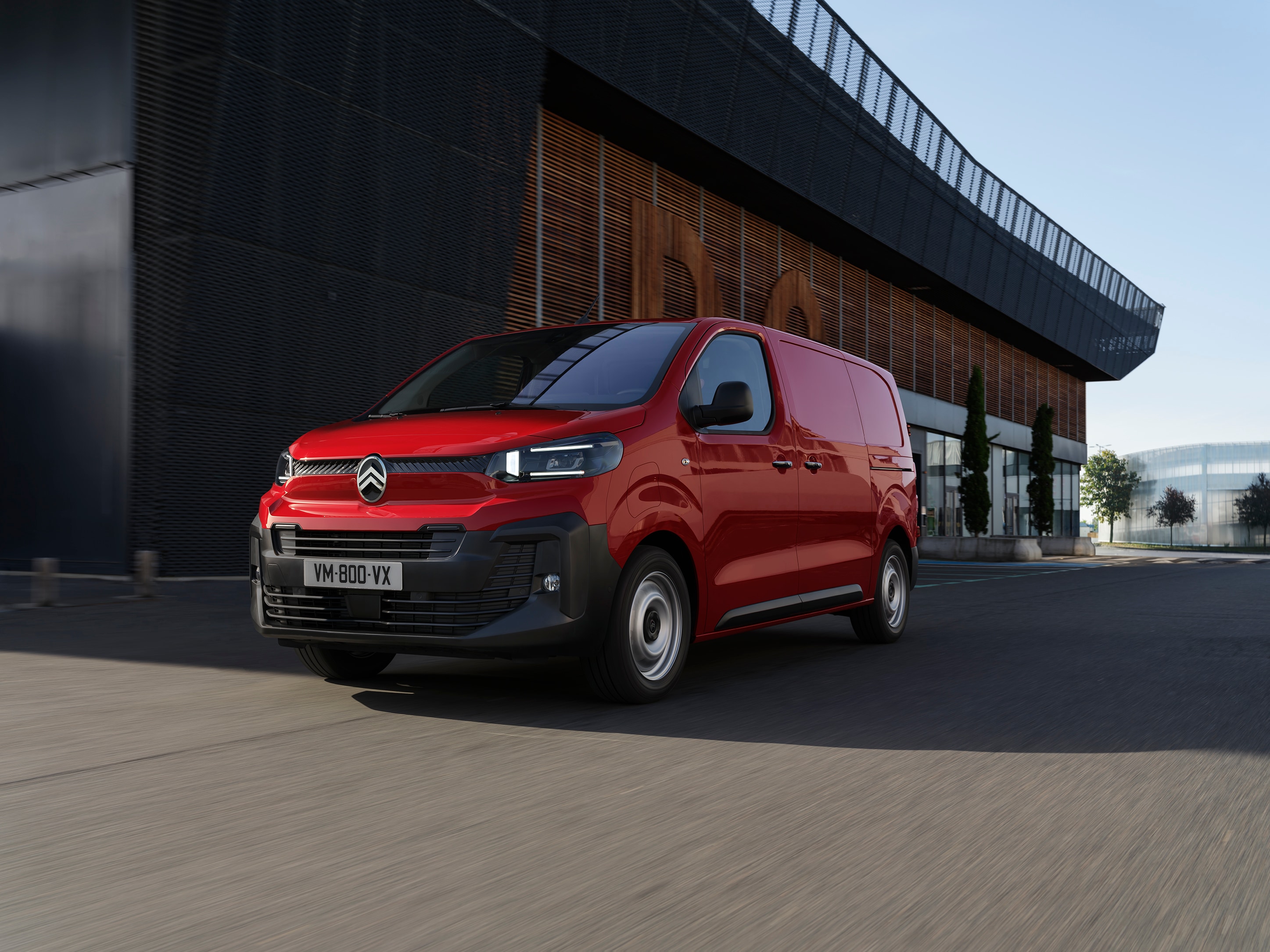 Driekwart vooraanzicht van een rode Citroën Jumpy geparkeerd bij een modern gebouw, met een professioneel design, praktische inzetbaarheid en een zelfverzekerde uitstraling.