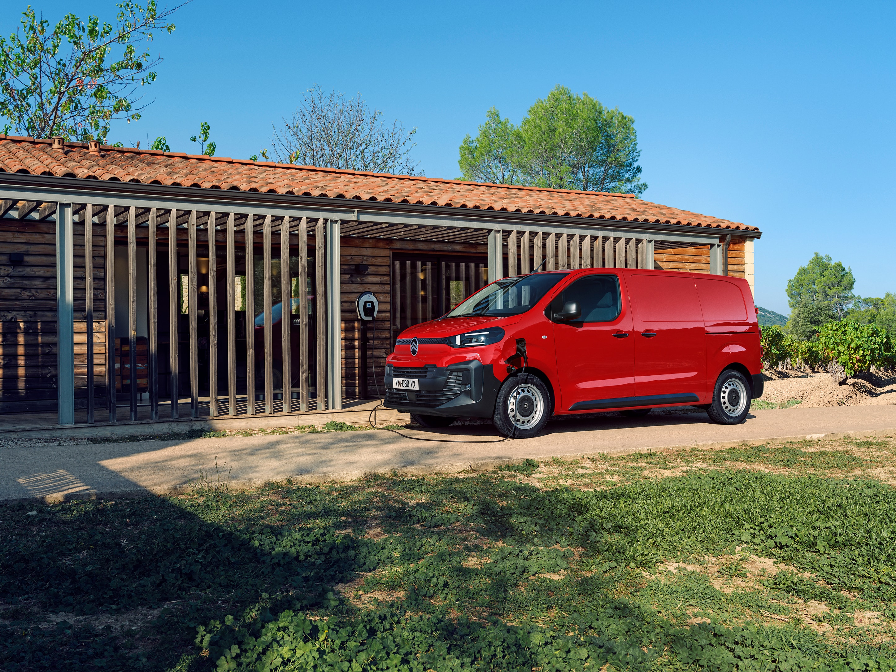 Zijaanzicht in driekwart perspectief van een rode Citroën ë‑Jumpy die wordt opgeladen voor een gebouw, met focus op elektrische mobiliteit, dagelijks gebruiksgemak en een zelfverzekerde professionele uitstraling.