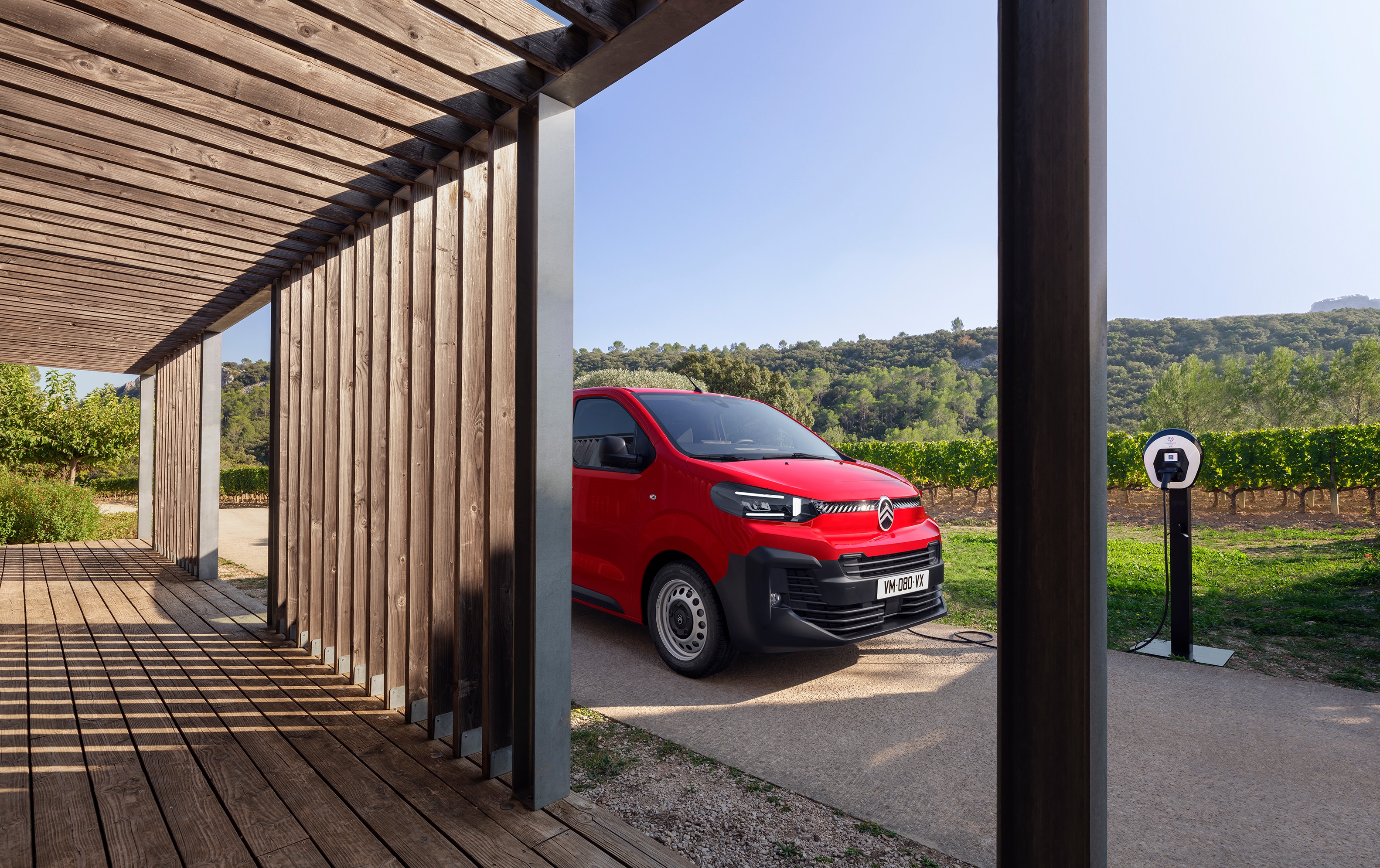 Zijaanzicht in driekwart perspectief van een rode Citroën ë‑Jumpy geparkeerd bij een houten gebouw, met een modern elektrisch design, praktische inzetbaarheid en een zelfverzekerde professionele uitstraling.