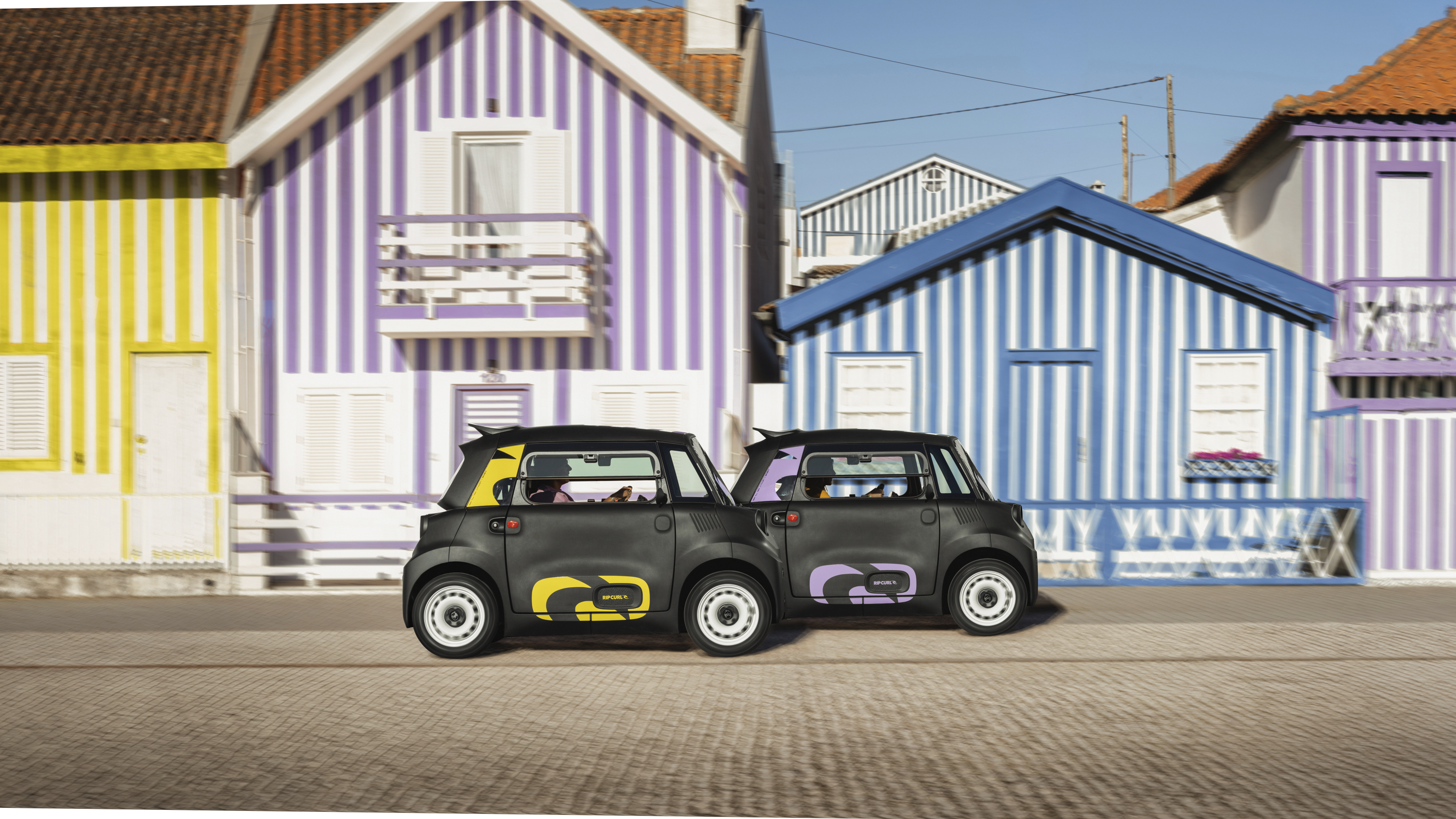Zijaanzicht van twee Citroën Ami x Rip Curl‑modellen naast elkaar in een straat, met herkenbare stedelijke stijl.