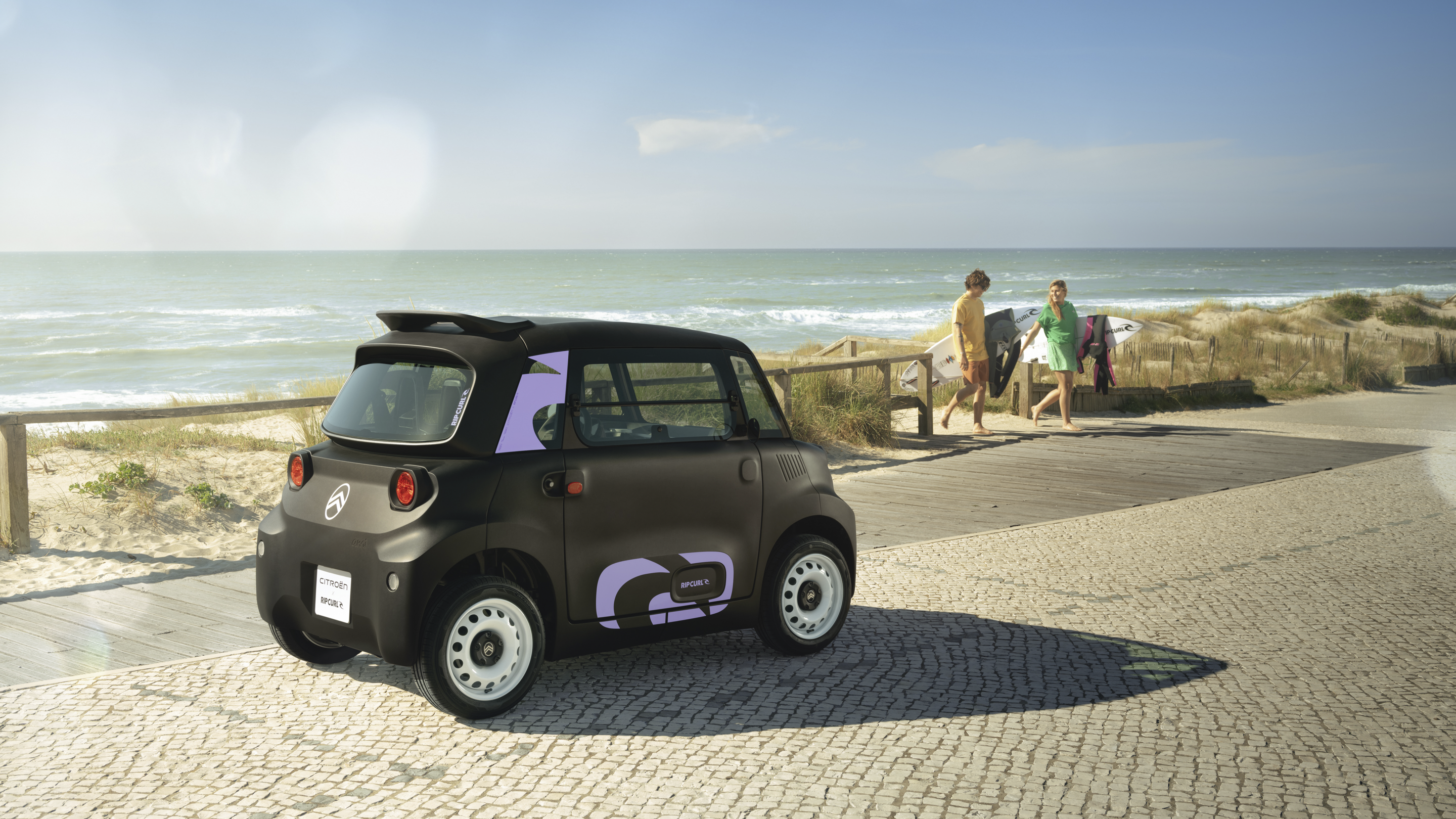 Driekwart achteraanzicht van de Citroën Ami x Rip Curl, elektrische auto zonder autorijbewijs, met kustachtige setting en stedelijke mobiliteit.