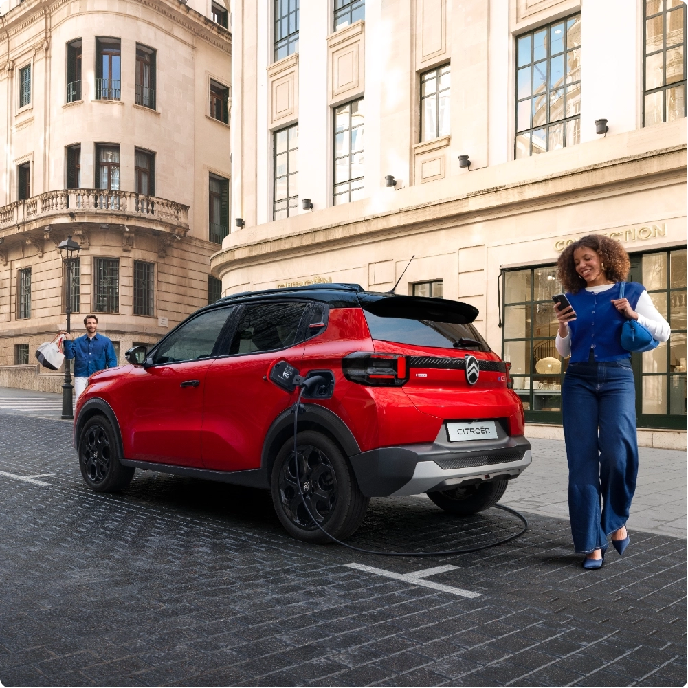 Driekwart achteraanzicht van de Citroën ë‑C3 elektrische SUV aan het opladen in de straat, met nadruk op praktische toegang tot de bagageruimte voor comfortabele, efficiënte en betaalbare mobiliteit.