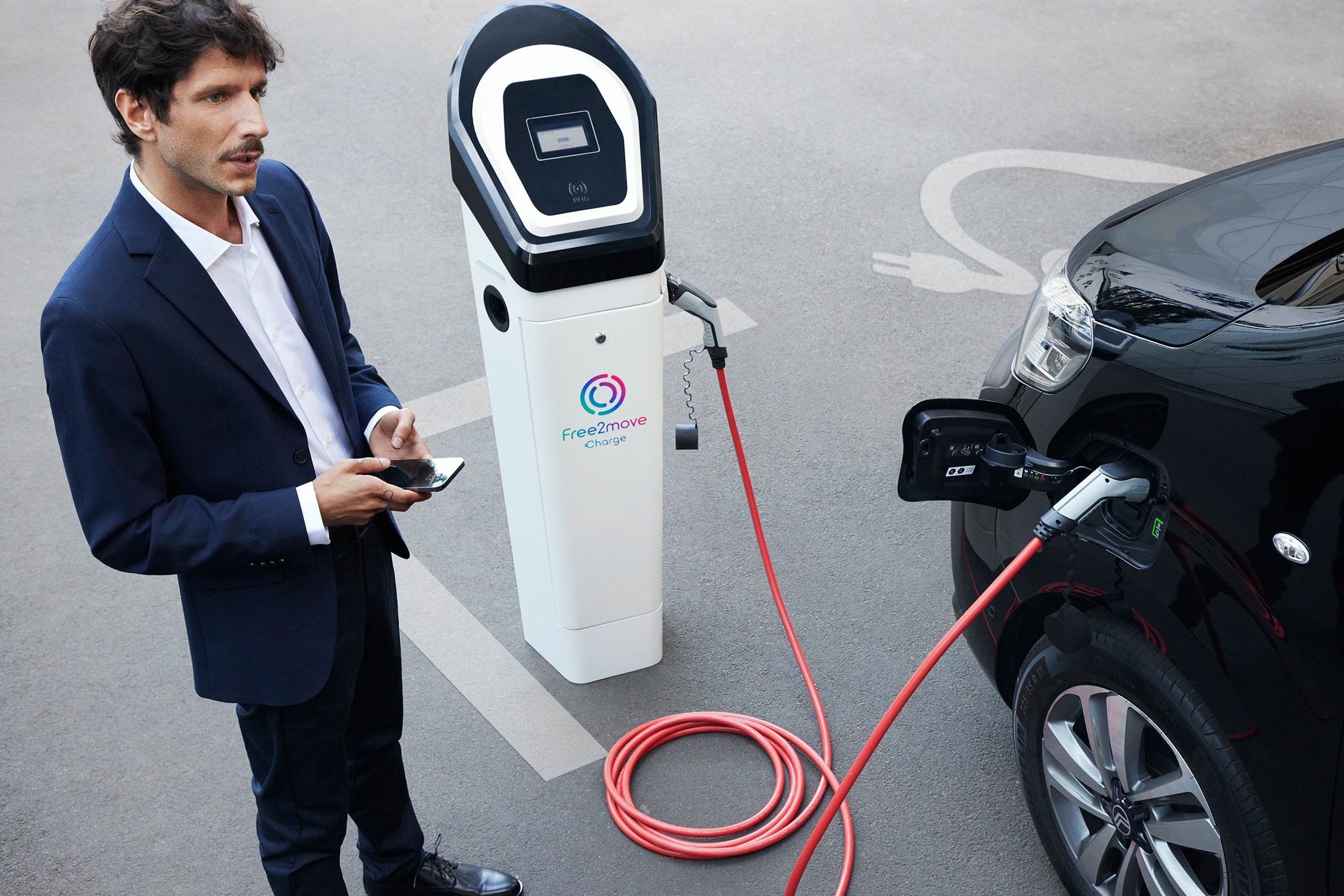 Elektrisch voertuig aan het opladen bij een openbaar laadpunt terwijl een smartphone‑app wordt gebruikt voor laadbeheer met Free2move Charge.